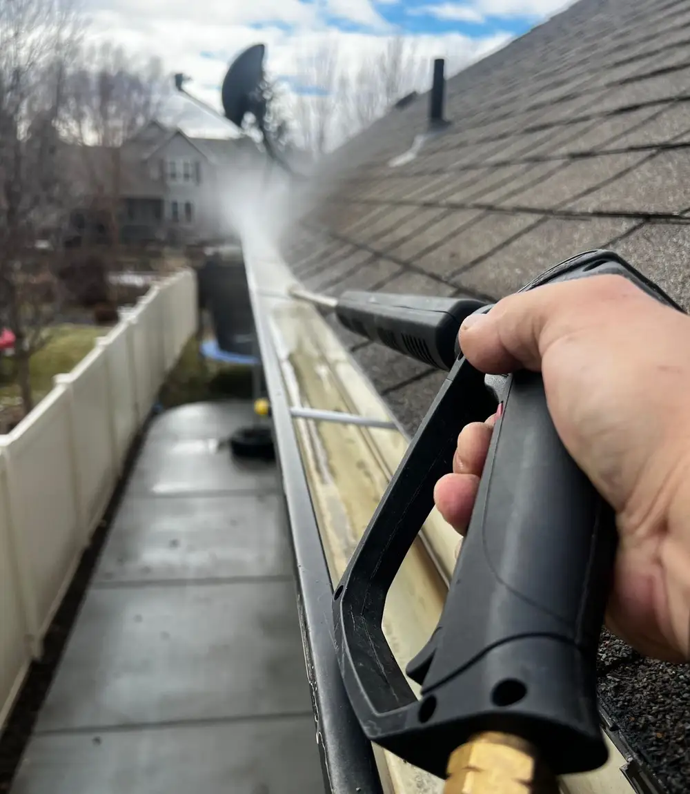 Hand holding a pressure-washer gun cleaning a gutter under a blue sky